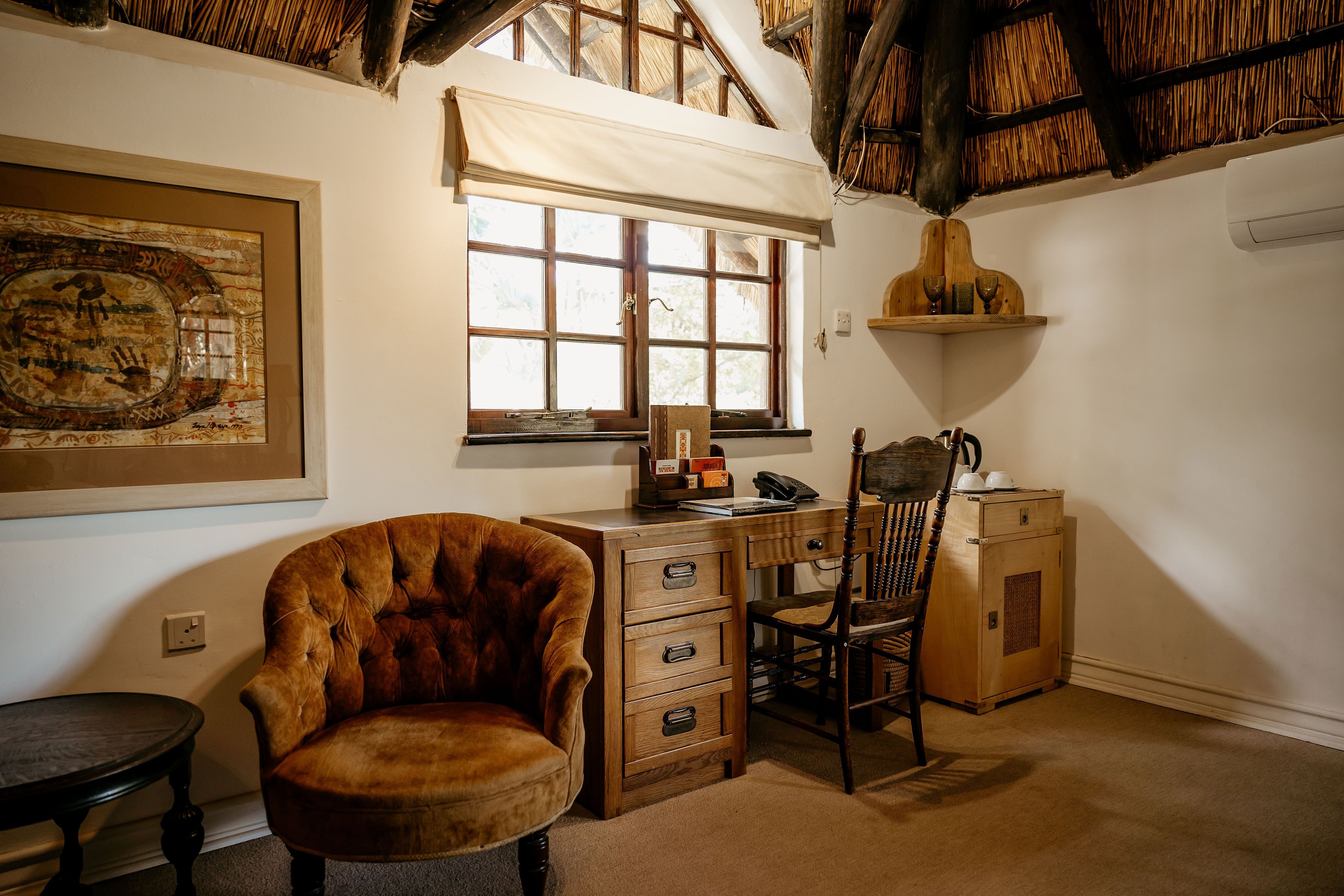 A room with a wooden desk, a chair, a framed artwork, a window with a white blind, and a thatched ceiling.