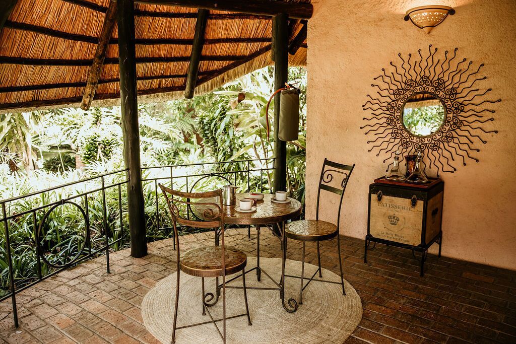 A covered outdoor seating area with a table and chairs, surrounded by a metal railing and lush greenery.