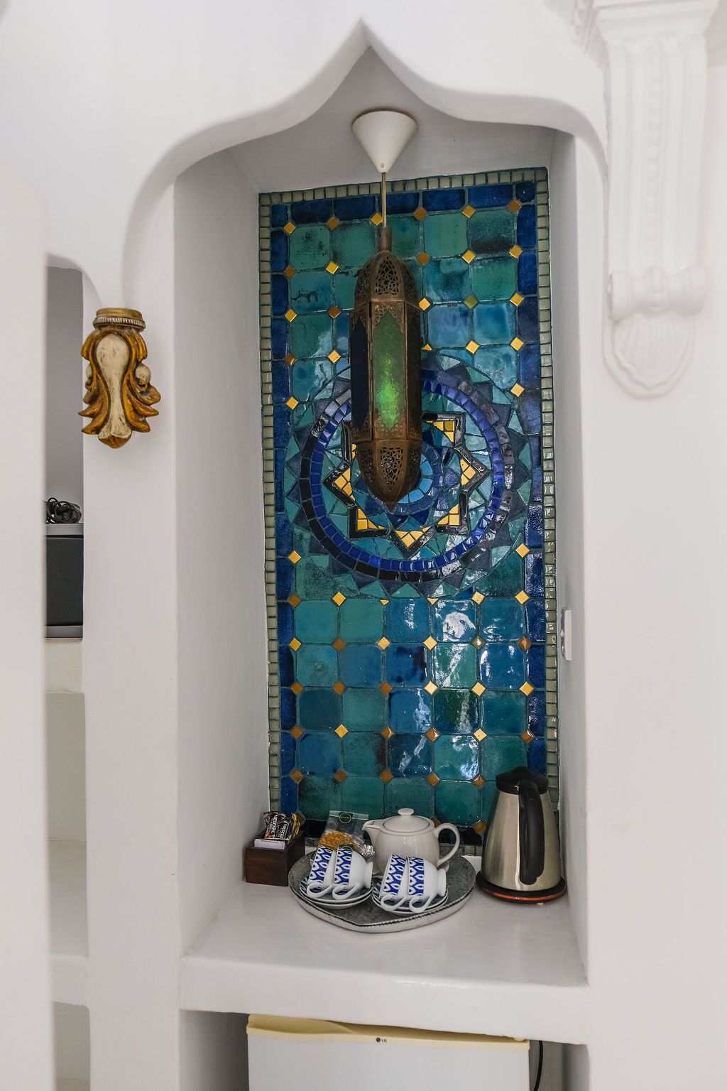 A room with a decorative tile wall, a small shelf with a teapot, cups, and a kettle.