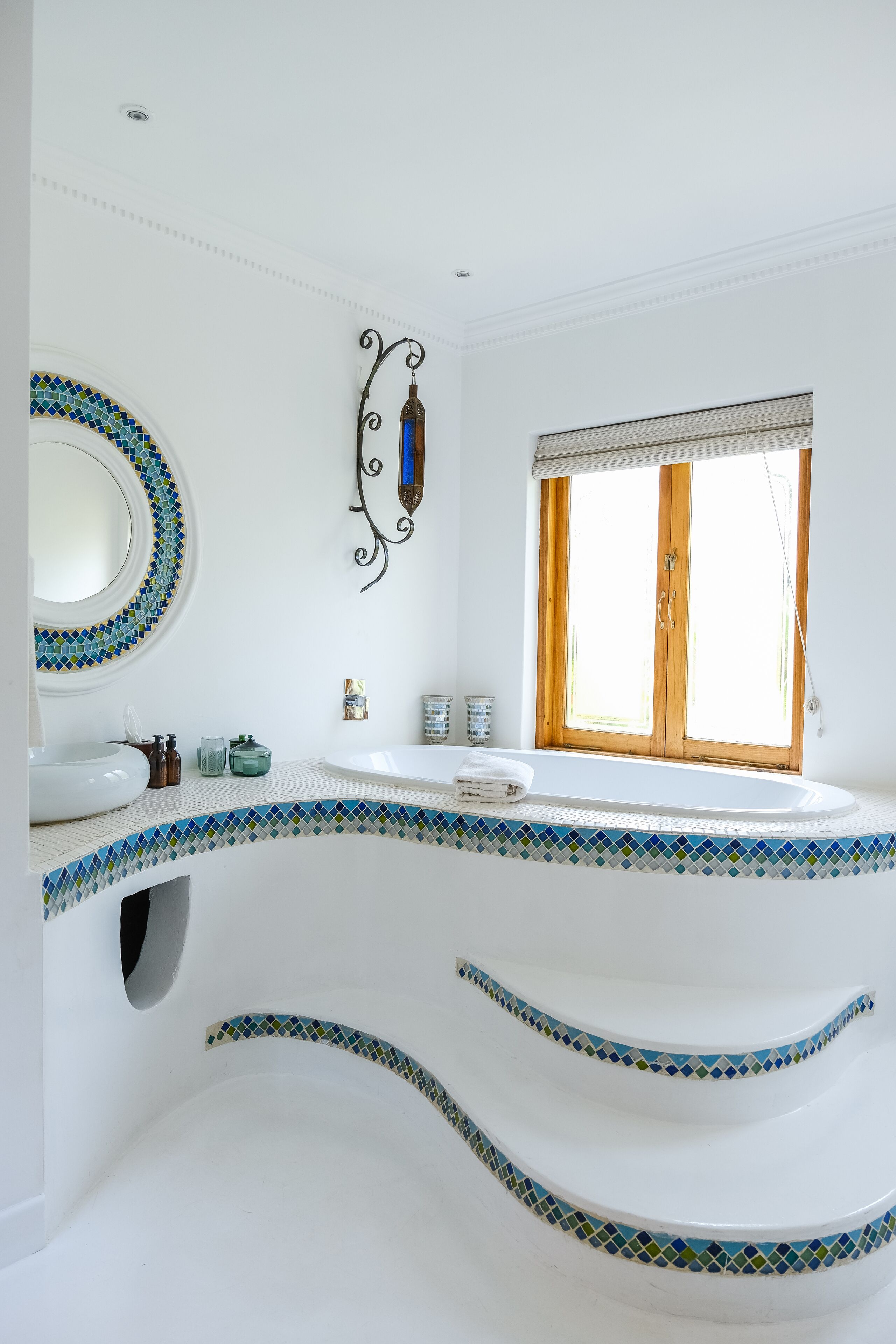 Ein Badezimmer mit einem weißen Waschbecken, einem Spiegel mit Mosaikrahmen, einer Dusche mit Holztür und einem Fenster mit Jalousien.