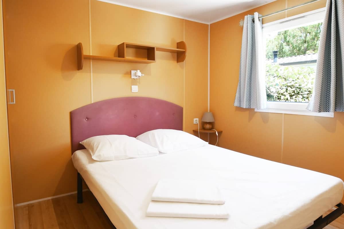 A bedroom with a single bed, wooden flooring, a headboard, a bedside table with a lamp, and a window with curtains.