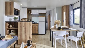 A compact kitchen with wooden cabinets, a microwave, and a refrigerator. There is a dining area with a table and chairs, and a window with curtains. - Camping Les Castors (Calvi)