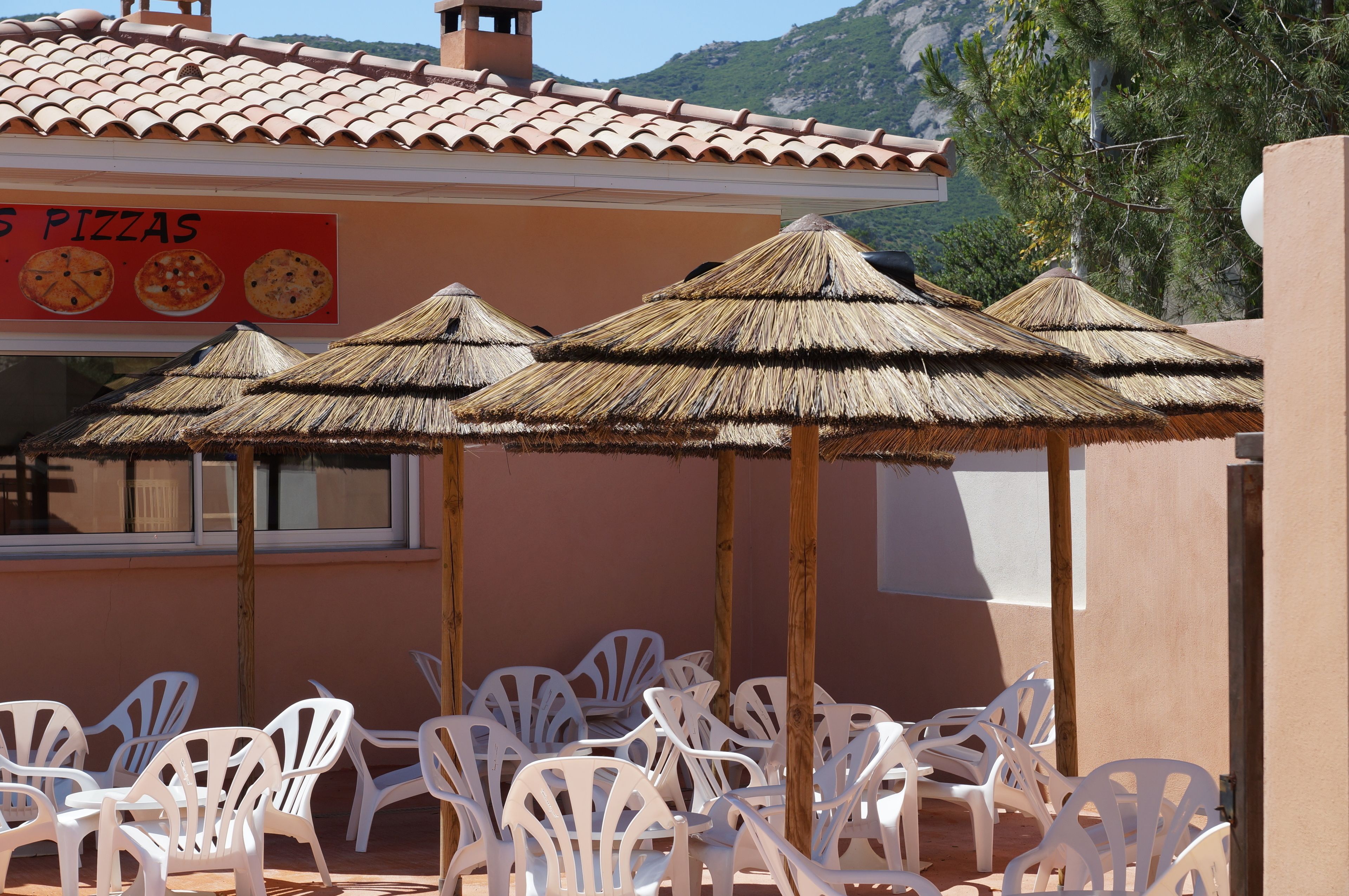 A restaurant with outdoor seating, thatched umbrellas, and a sign advertising pizzas.