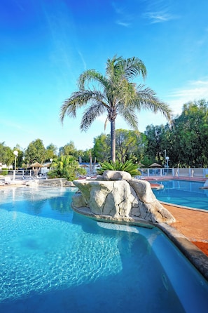 A clear blue pool with a palm tree and rock formations, surrounded by lounge chairs and umbrellas. - Camping Les Castors (Calvi)