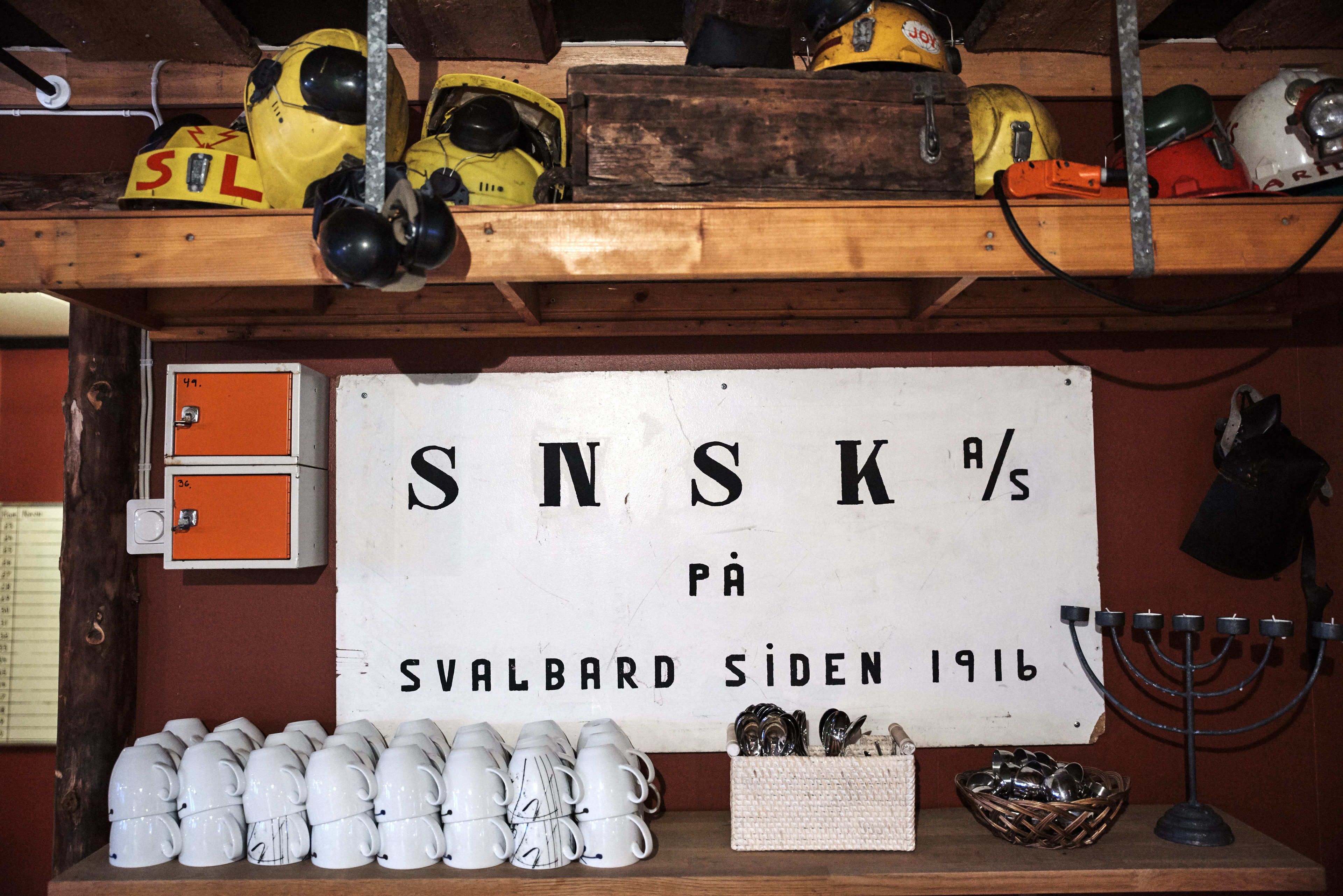 A room with a sign reading "SNSK %s på Svalbard Siden 1916" and a collection of white mugs on the floor.