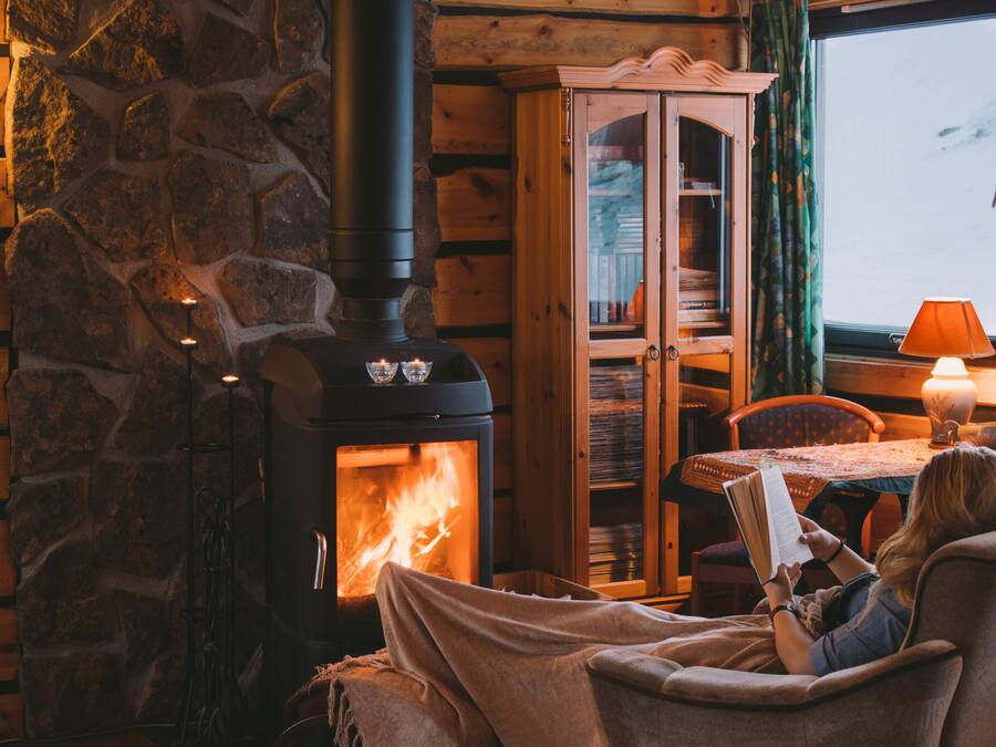 A cozy cabin interior with a stone wall, a wood-burning stove, a wooden cabinet, and a person reading by a table with a lamp.