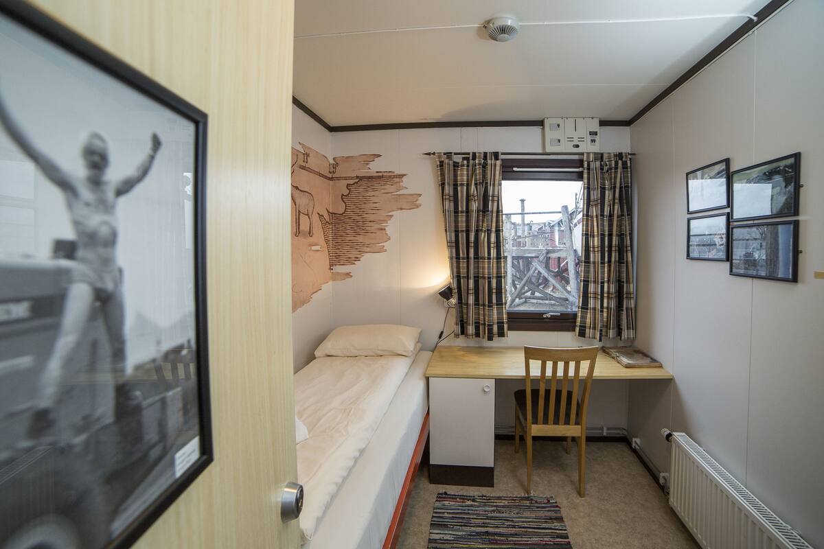 A compact bedroom with a single bed, a desk, and a chair. There is a window with curtains, a radiator, and a wall-mounted light.