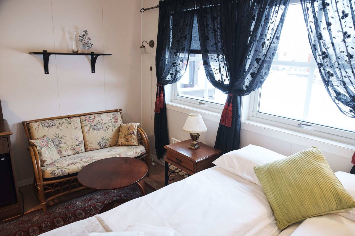 A room with a bed, a sofa, a small table, a lamp, and a window with curtains.