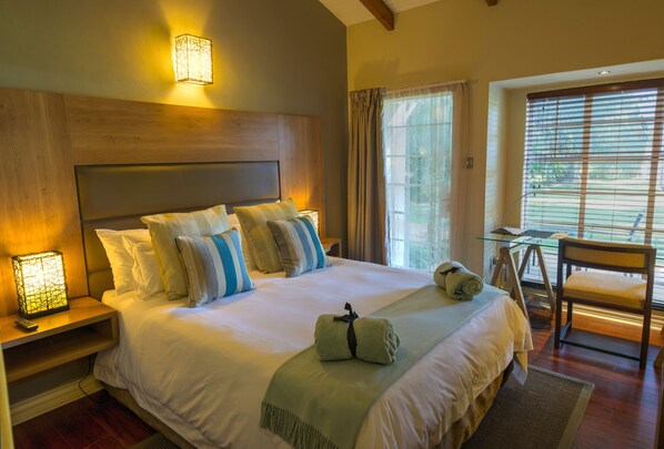 A large bed with multiple pillows, a bedside table with a lamp, a chair, and a window with curtains. - Sandalwood Lodge (Harare)