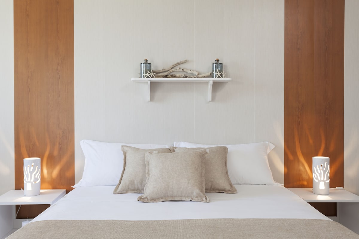 Quarto com cama, duas mesas de cabeceira, prateleira com objetos decorativos e iluminação na parede.