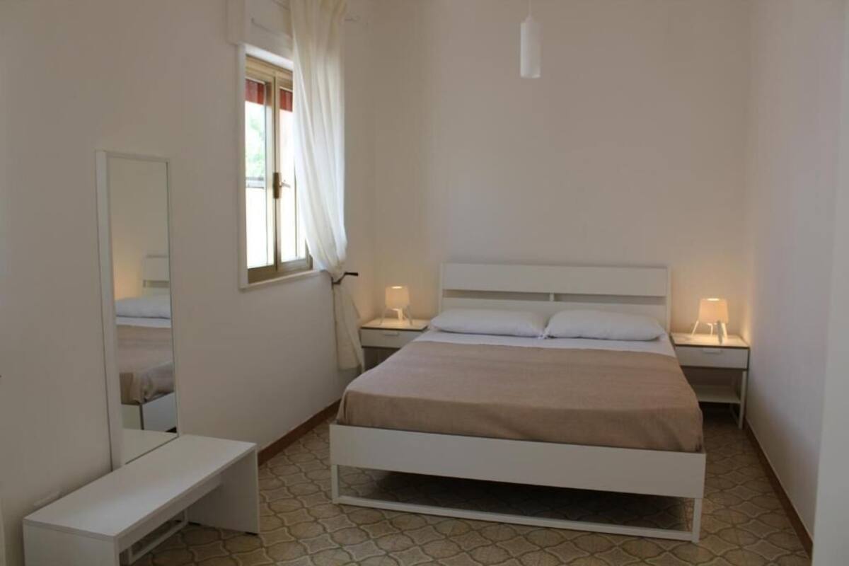 Quarto com uma cama de solteiro, mesas de cabeceira, um espelho e uma janela com cortinas.