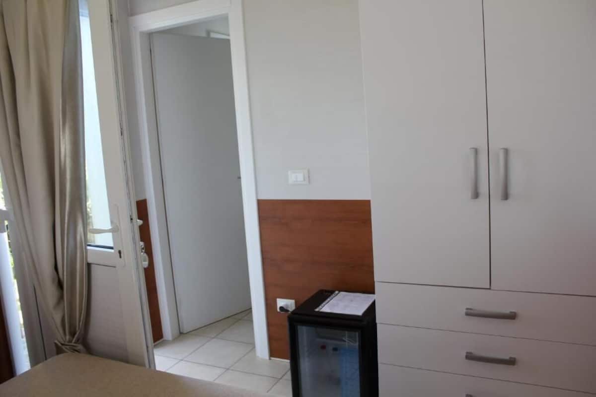 Quarto de hotel com cama, guarda-roupa, secretária com computador e janela com cortinas.