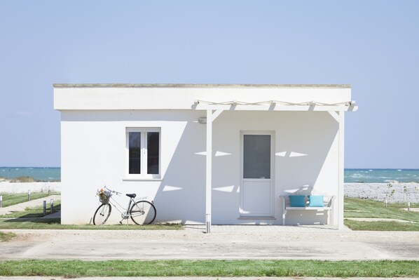 A small white building with a single door and window, a bicycle parked outside, and a bench with a cushion.