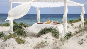 Un paisaje de playa con una toldo blanco y una mesa dispuesta con comida y bebidas.