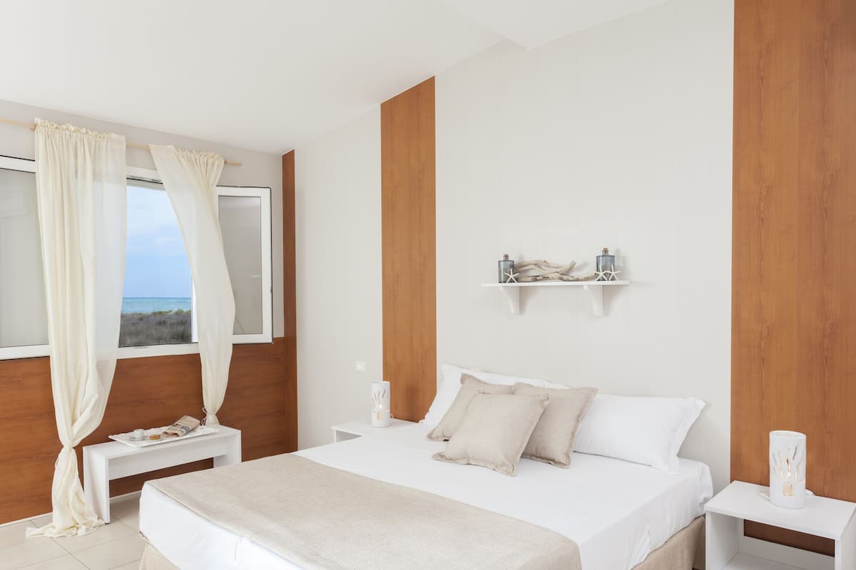 Quarto com uma cama grande, mesa de cabeceira, janela com cortinas e vista para o mar.