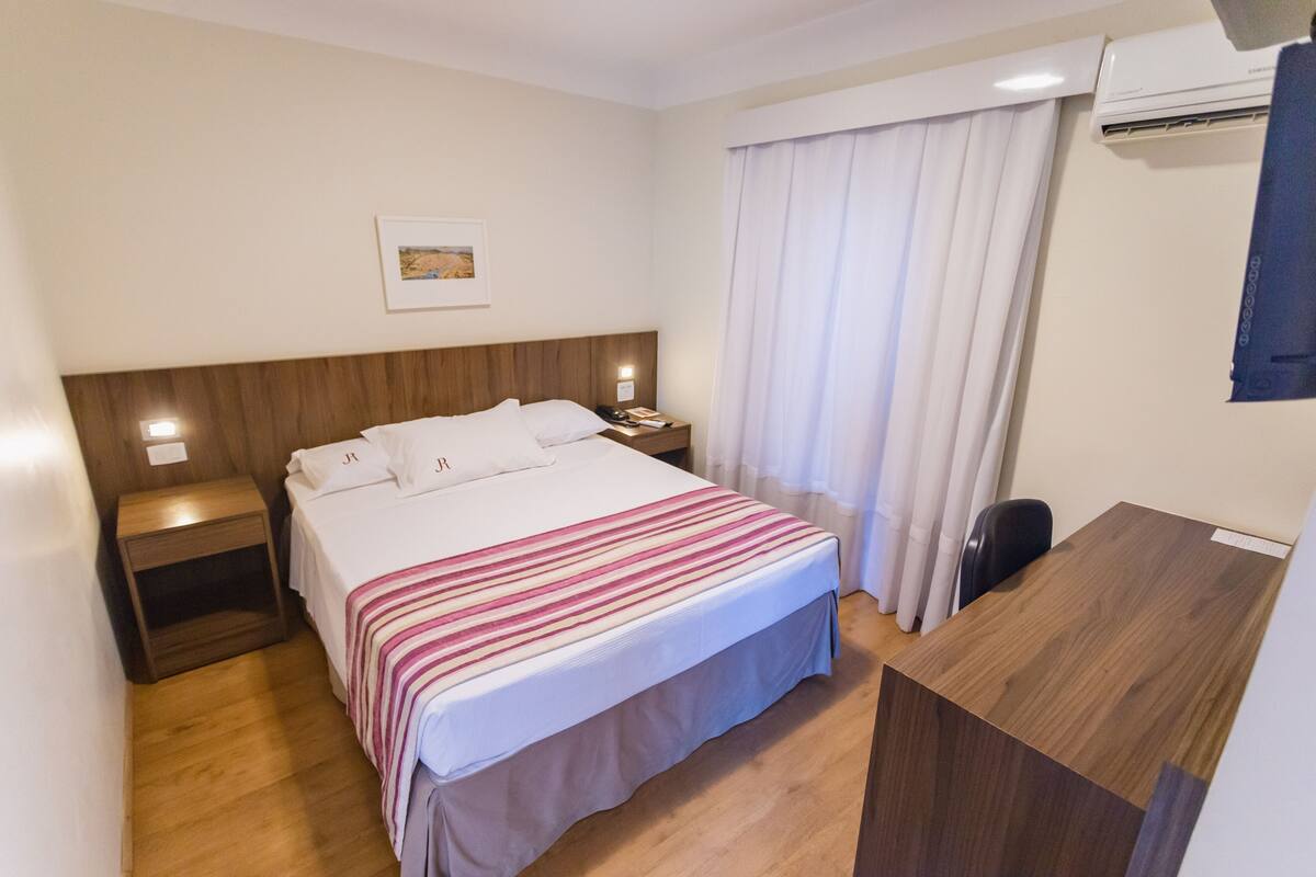 Una camera d'albergo con un letto, un comodino, una scrivania, una sedia e una finestra con tende.