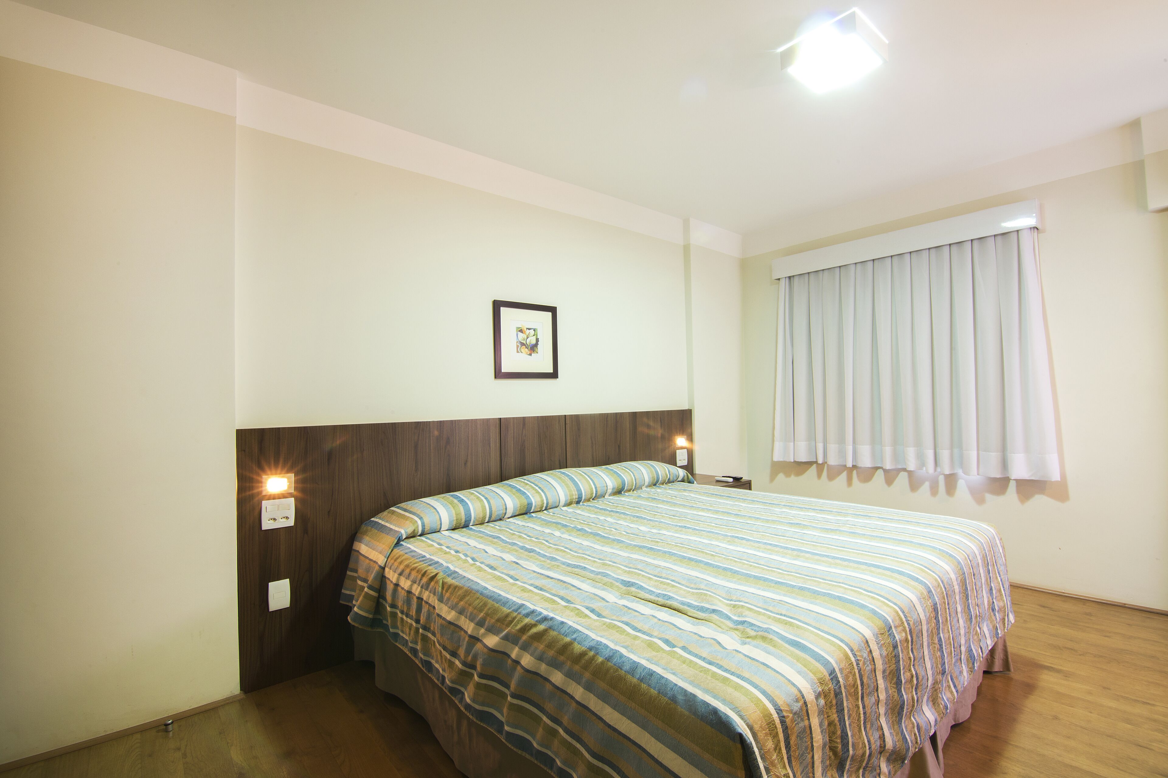 Habitación de hotel con cabecera de madera, cubrecama a rayas, un cuadro enmarcado en la pared y una ventana con cortinas.