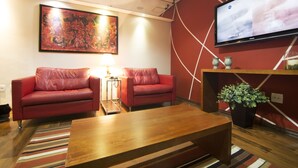 A modern living room with two red leather sofas, a wooden coffee table, a large abstract painting, a flat-screen TV, and a potted plant. - JR Hotel (Ribeirão Preto)