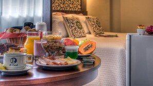 A breakfast buffet with various foods and drinks, a bed with patterned pillows, and a small potted plant. - JR Hotel (Ribeirão Preto)