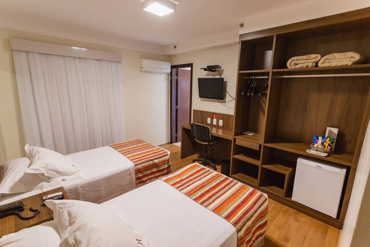 Camera d'albergo con due letti, una scrivania, una sedia, una TV, un mini-frigorifero e un armadio.