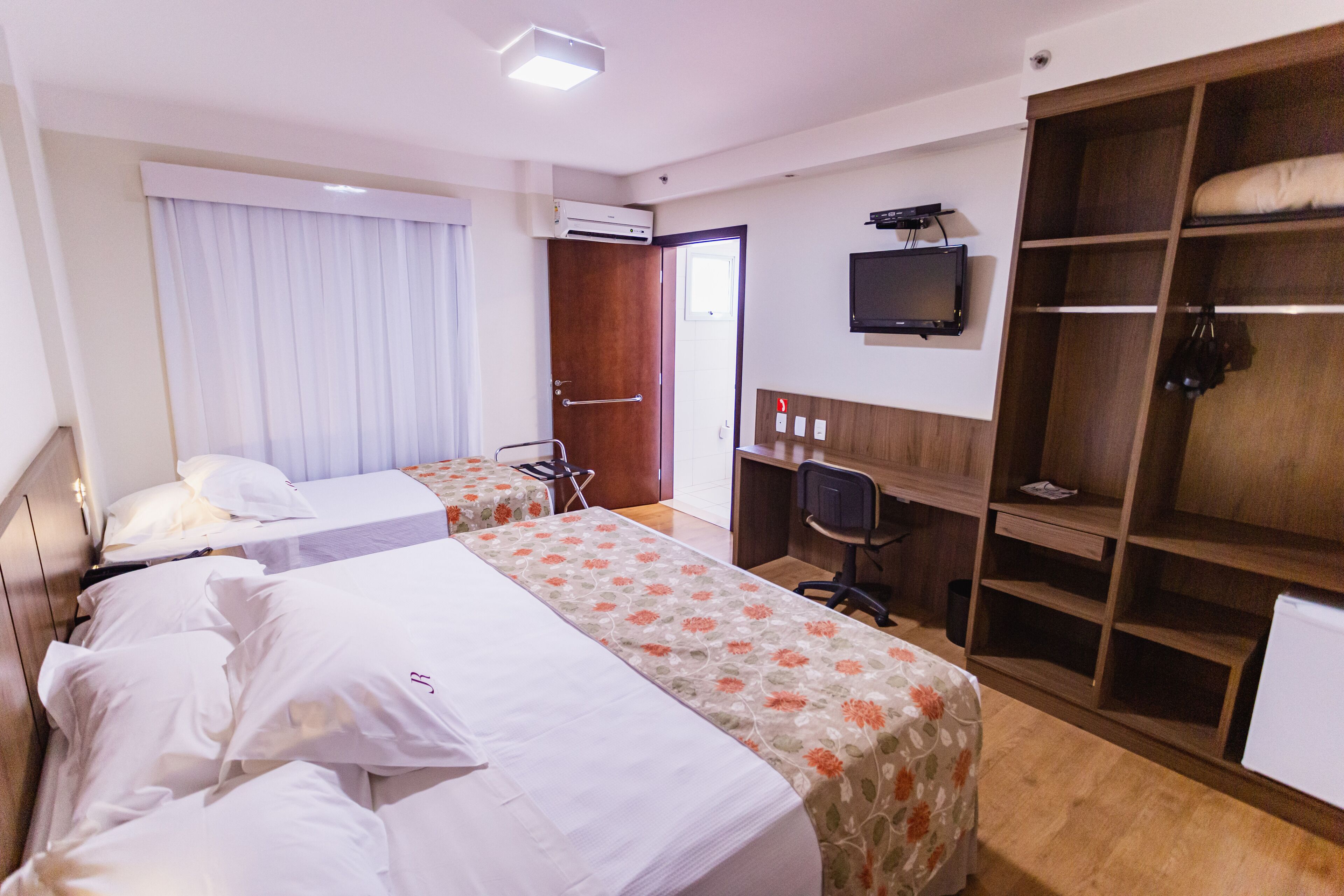 Quarto de hotel com duas camas, uma escrivaninha, um guarda-roupa e uma televisão.