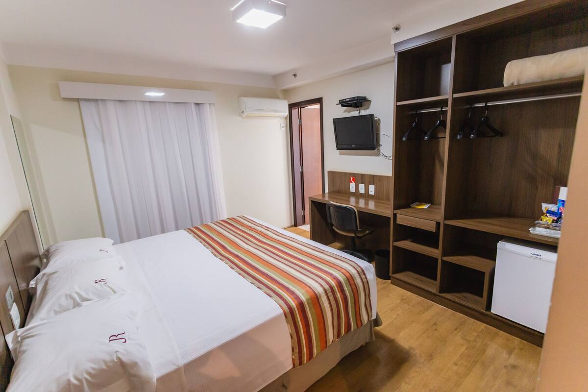 Camera d'hotel con un letto, una scrivania, una sedia, una TV, un armadio e un mini-frigorifero.