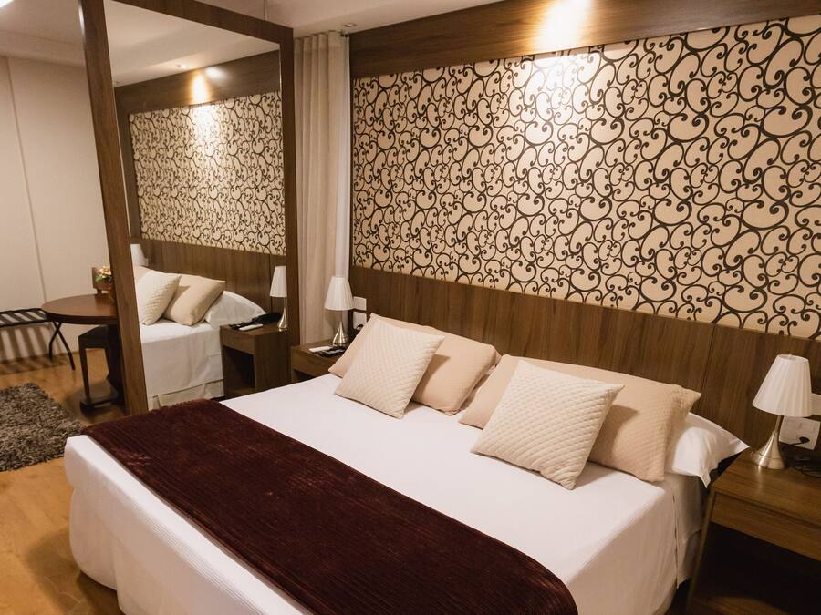 Cama bem arrumada com manta escura, cabeceira de madeira e papel de parede estampado.
