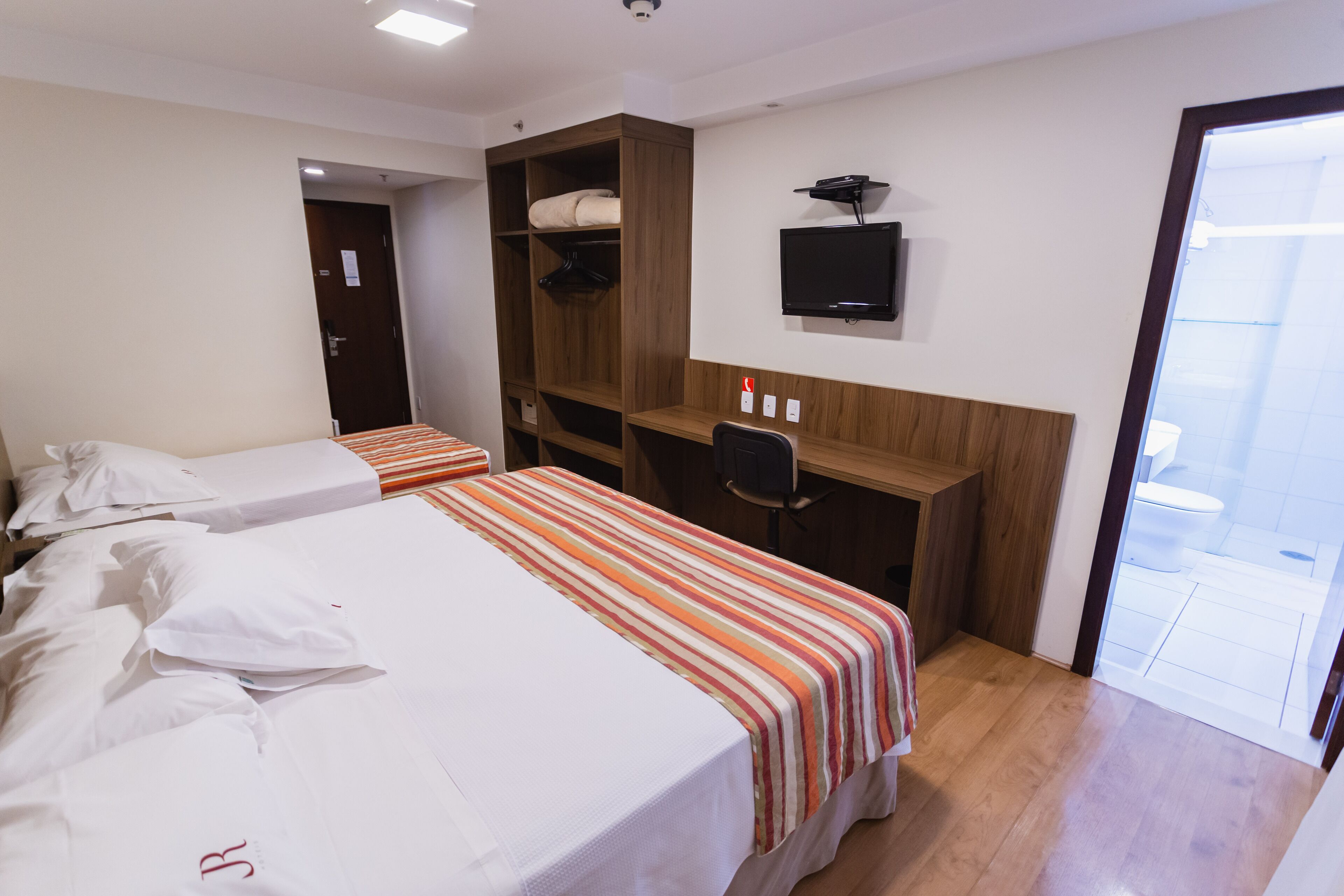 Ein Hotelzimmer mit zwei Betten, einem Holzschrank, einem Schreibtisch mit Stuhl, einem Fernseher und einer Glastür zum Balkon.