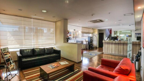 A hotel lobby with a black leather sofa, a red armchair, and a wooden coffee table. There is a reception desk in the background with a counter and a person behind it. - JR Hotel (Ribeirão Preto)
