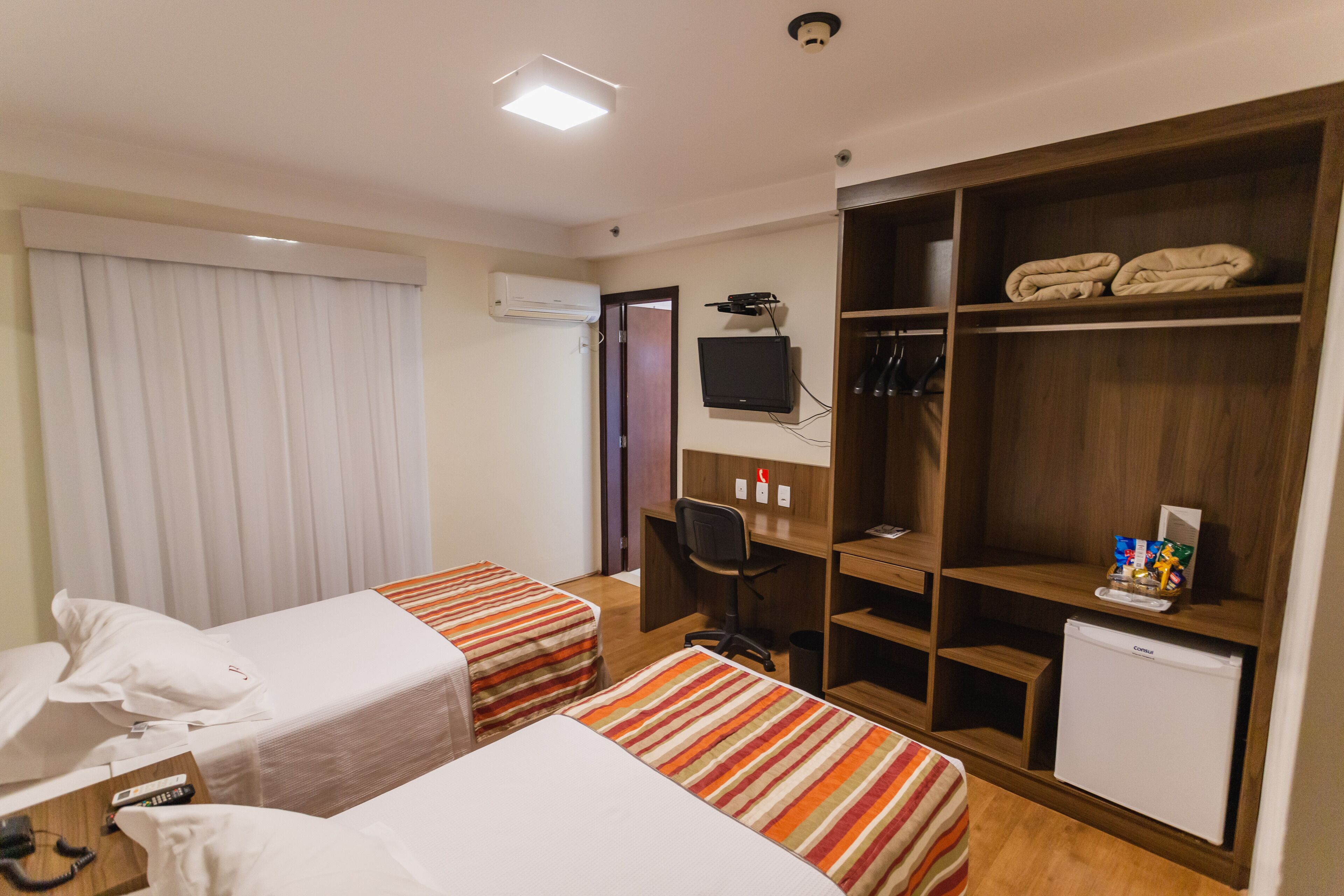 Habitación de hotel con dos camas, un escritorio, una silla, un televisor, un armario y un refrigerador pequeño.