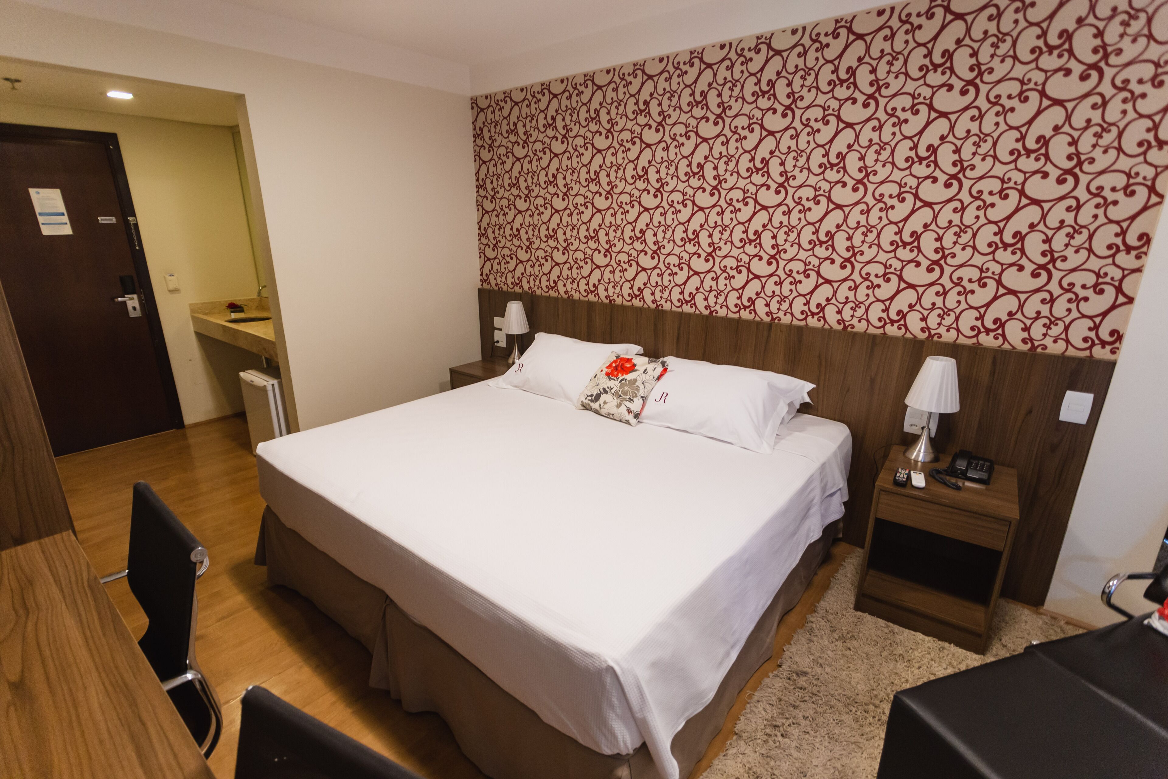 Una habitación de hotel con una cama grande, un escritorio de madera, una silla, una mesita de noche con lámpara y un papel tapiz estampado.