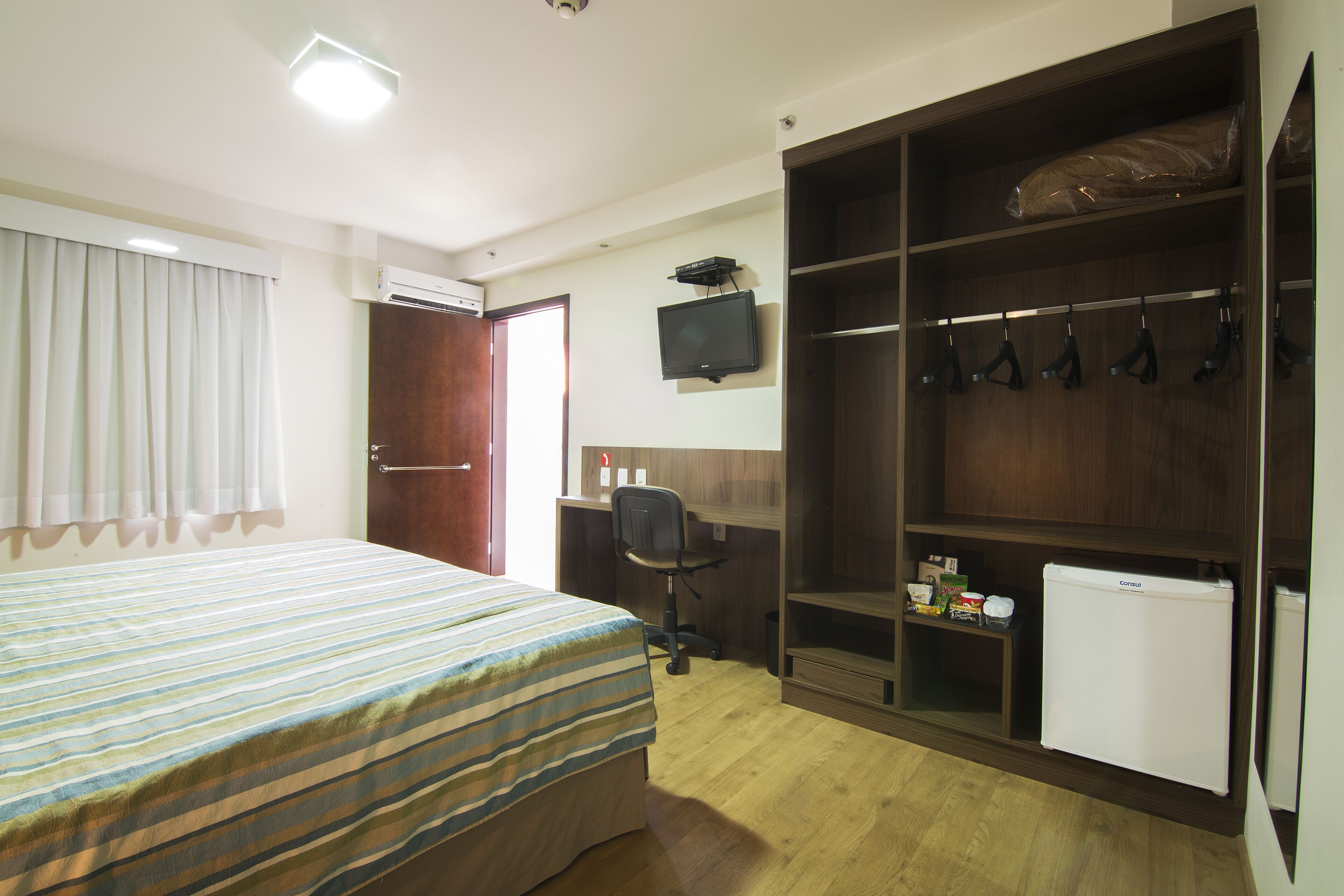 Ein Hotelzimmer mit Bett, Schreibtisch, Stuhl, Fernseher, Kleiderschrank und einem kleinen Kühlschrank.