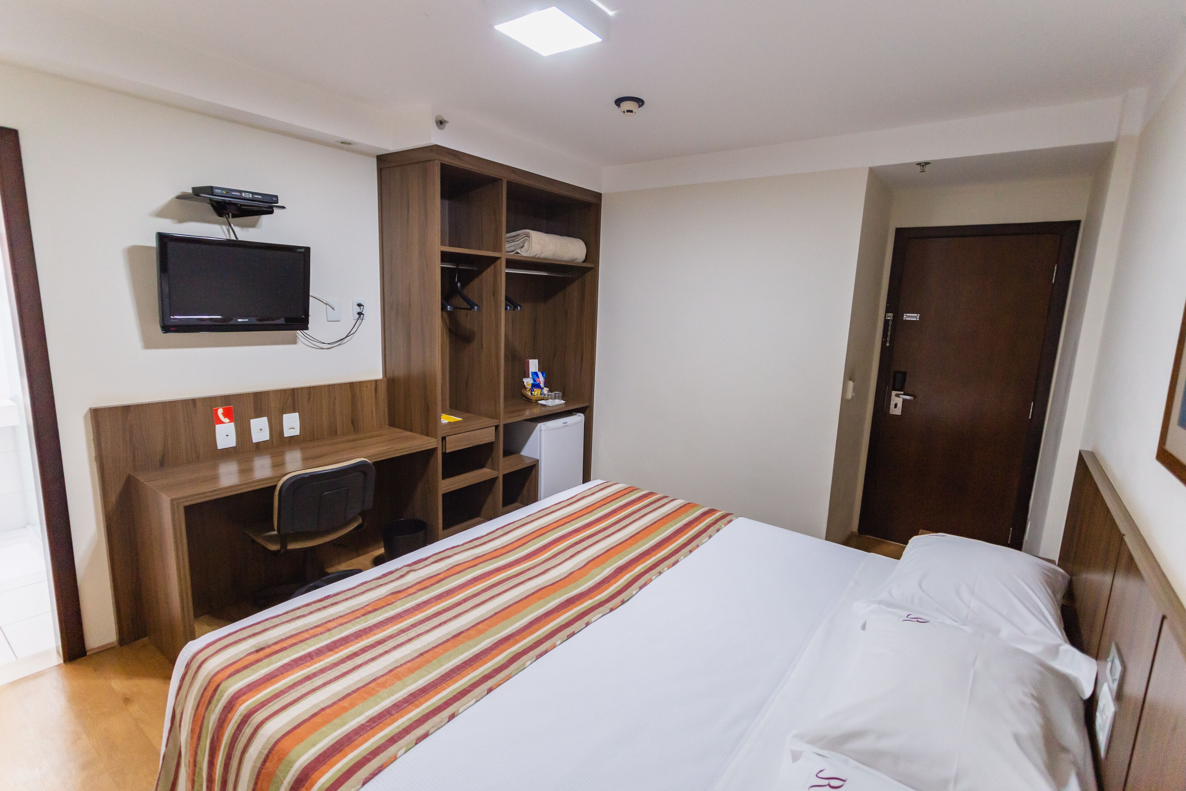 A hotel room with a bed, a desk with a chair, a television mounted on the wall, and a wardrobe.