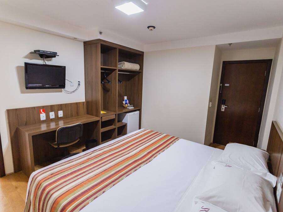 A hotel room with a bed, a desk with a chair, a television mounted on the wall, and a wardrobe.