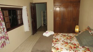A bedroom with a bed, a nightstand, a lamp, a wooden wardrobe, a window with curtains, and a door leading to another room. - Charitas Inn (Niteroi)