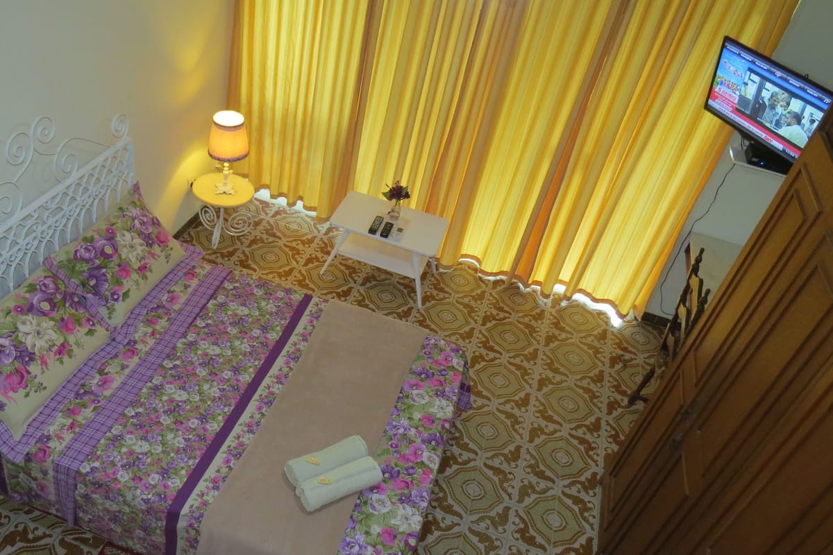 A bedroom with a bed, a bedside table with a lamp, a TV on the wall, and patterned curtains.