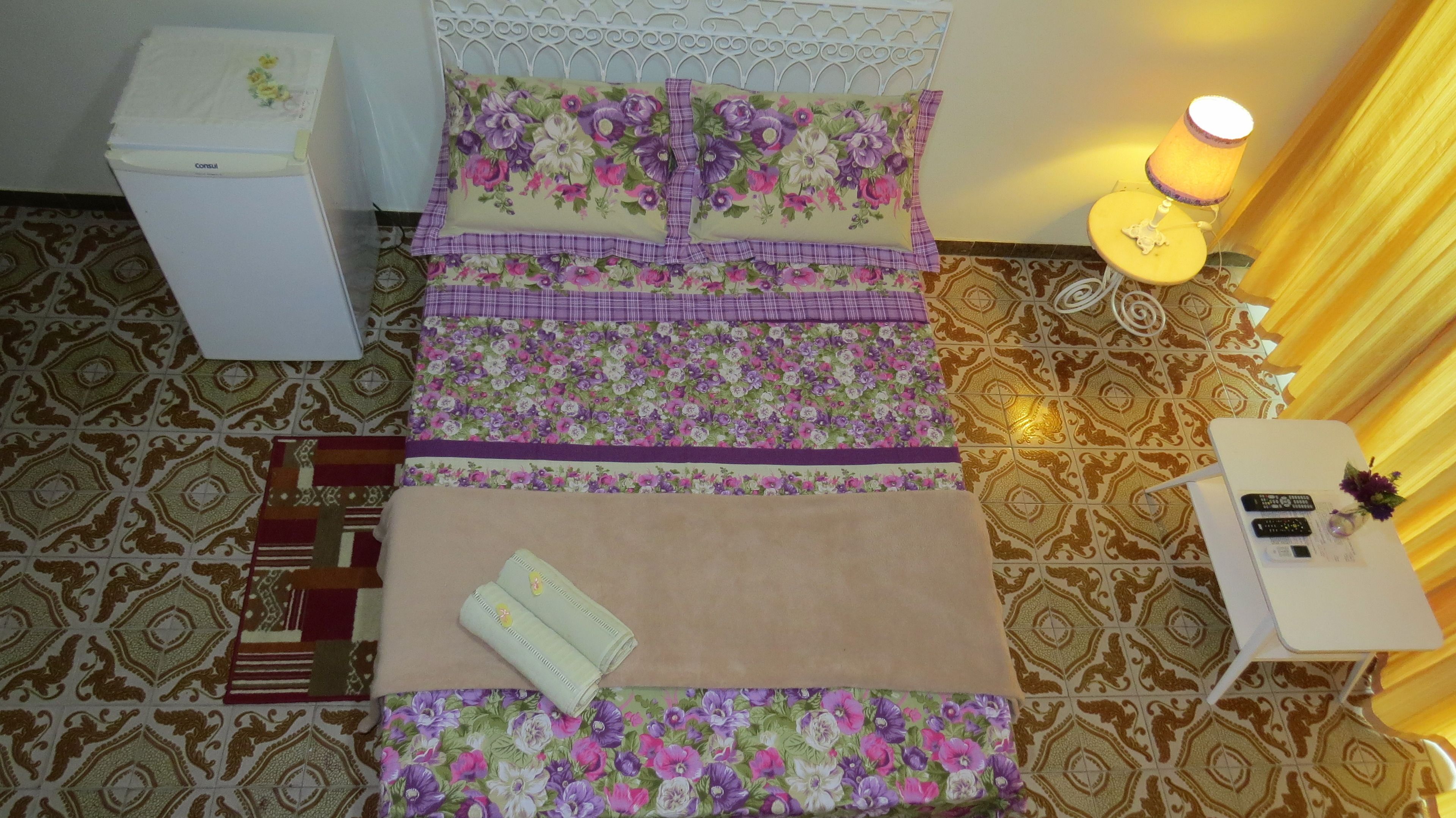 A neatly arranged room with a bed, bedside table, and a small table with a lamp and remote controls.