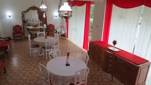 A dining area with a round table, white chairs, a wooden cabinet, and red curtains. - Charitas Inn (Niteroi)
