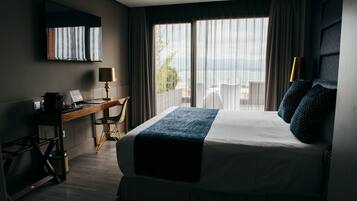 A hotel room with a large bed, a desk with a laptop, a chair, and a view of the outdoors through large windows.