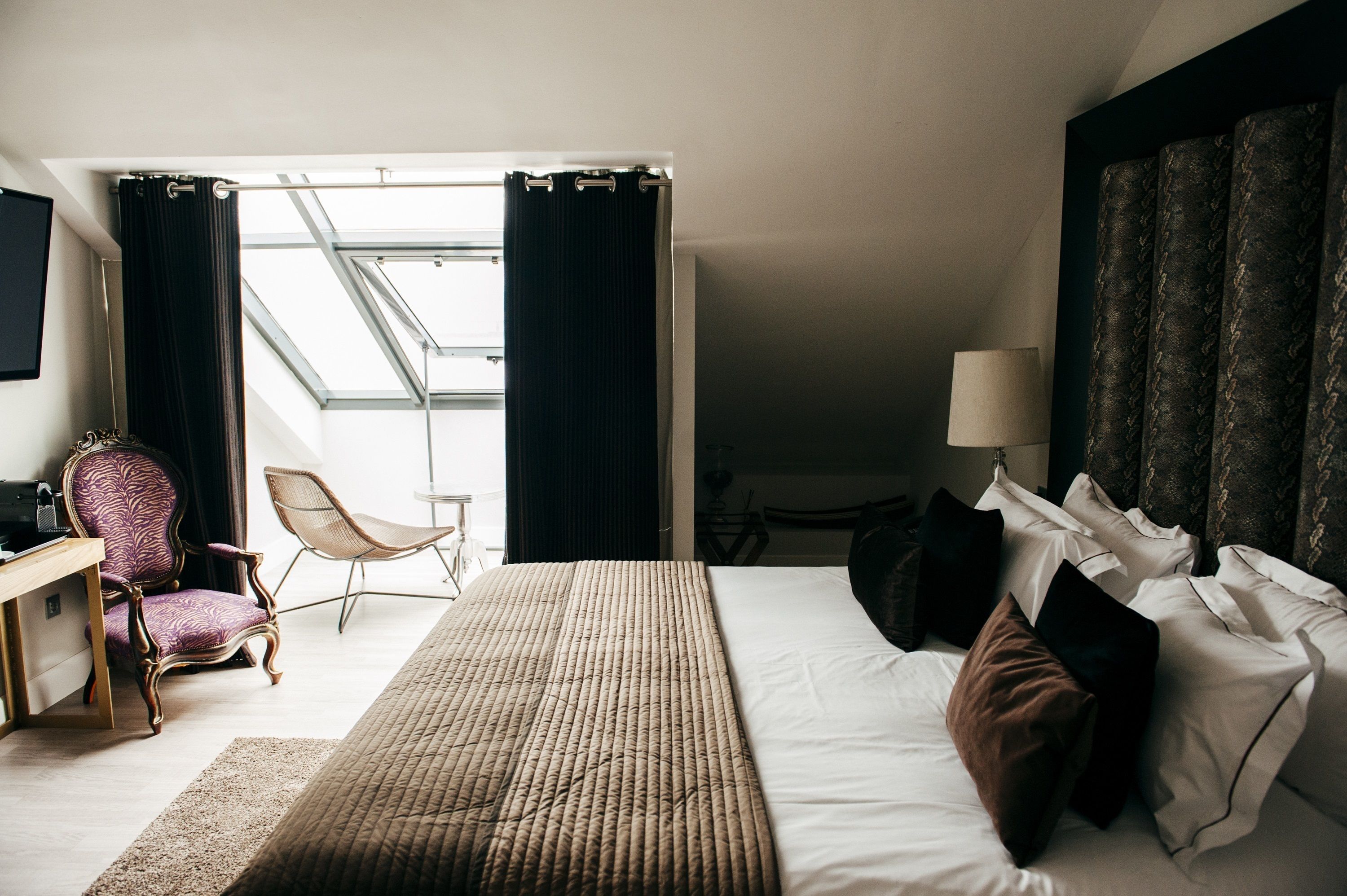 Une chambre d’hôtel avec un grand lit, un bureau avec une chaise, une fenêtre avec des rideaux et une lampe.