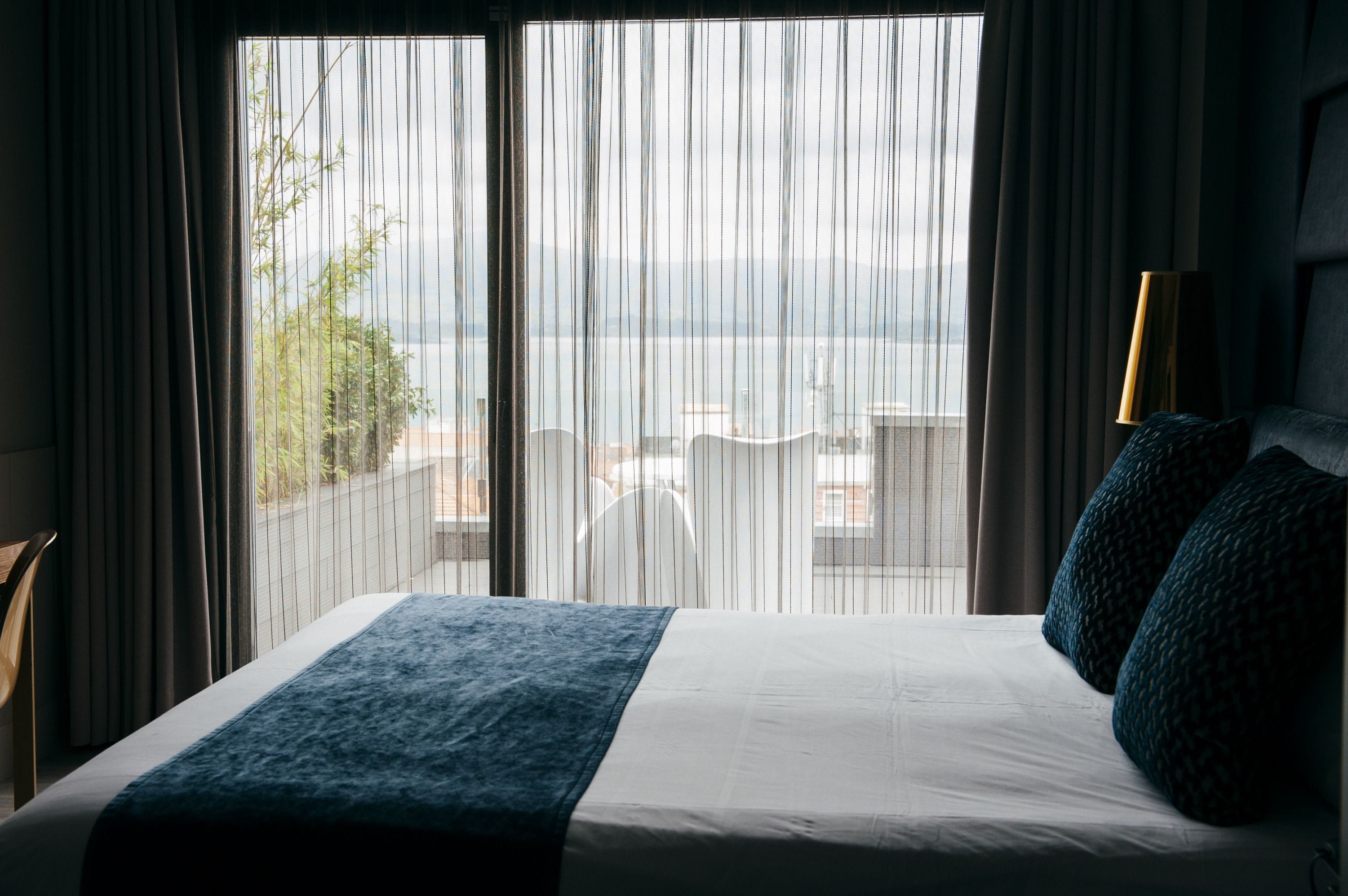 Een hotelkamer met een bed, uitzicht op de stad door dunne gordijnen, en een lamp op het nachtkastje.