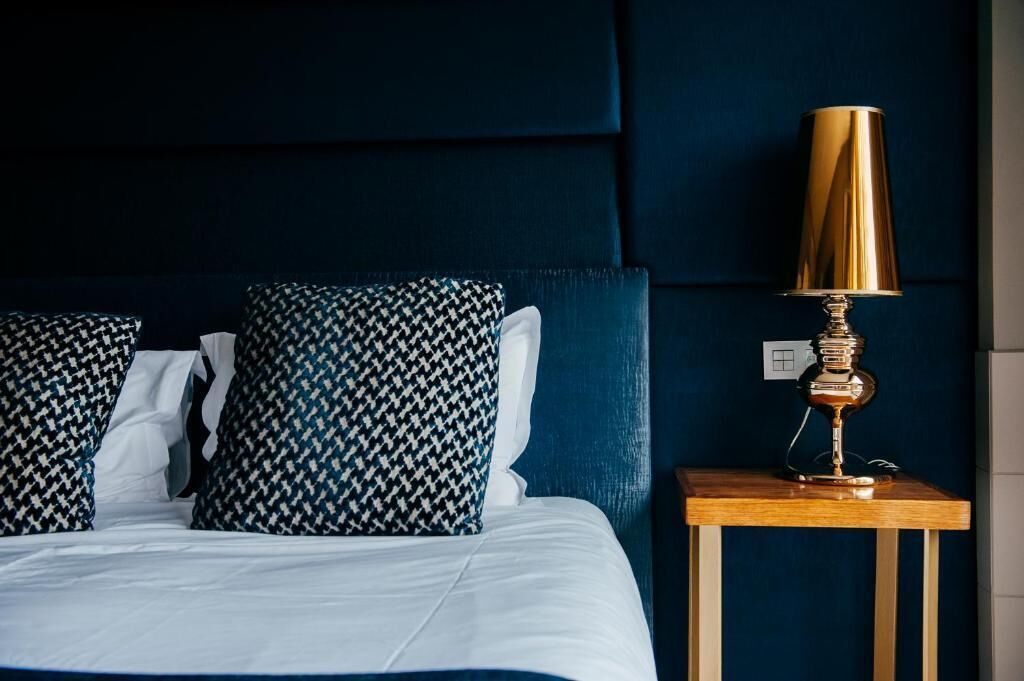 Una cama bien hecha con sábanas blancas, dos almohadones estampados, una lámpara dorada sobre una mesita de madera y un cabecero tapizado en azul oscuro.