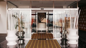 Entrada de hotel com porta de vidro, tapete estampado e dois vasos brancos com plantas.