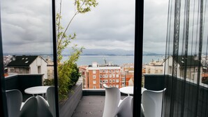 Uma varanda com cadeiras e mesa brancas, com vista para uma paisagem urbana e um corpo d'água.
