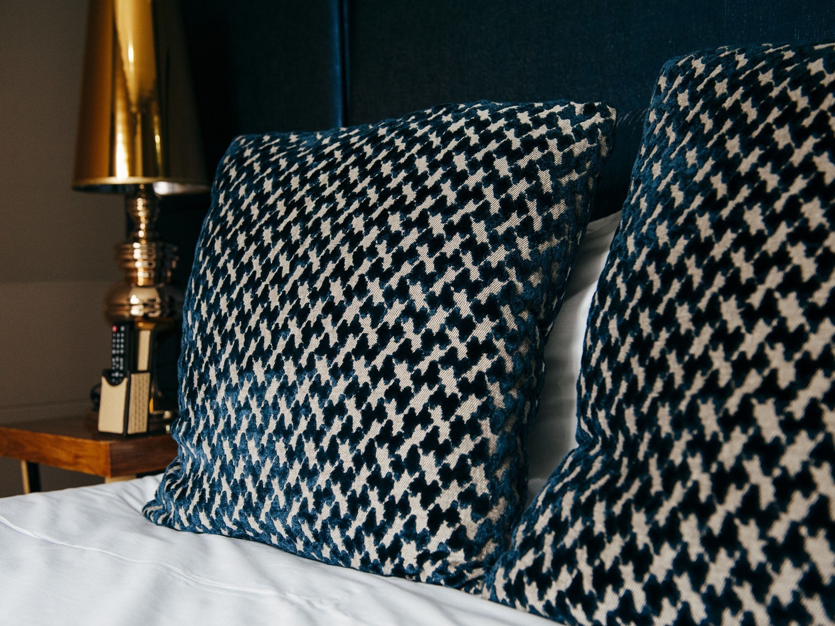 A bed with two houndstooth patterned pillows and a gold-colored lamp beside it.