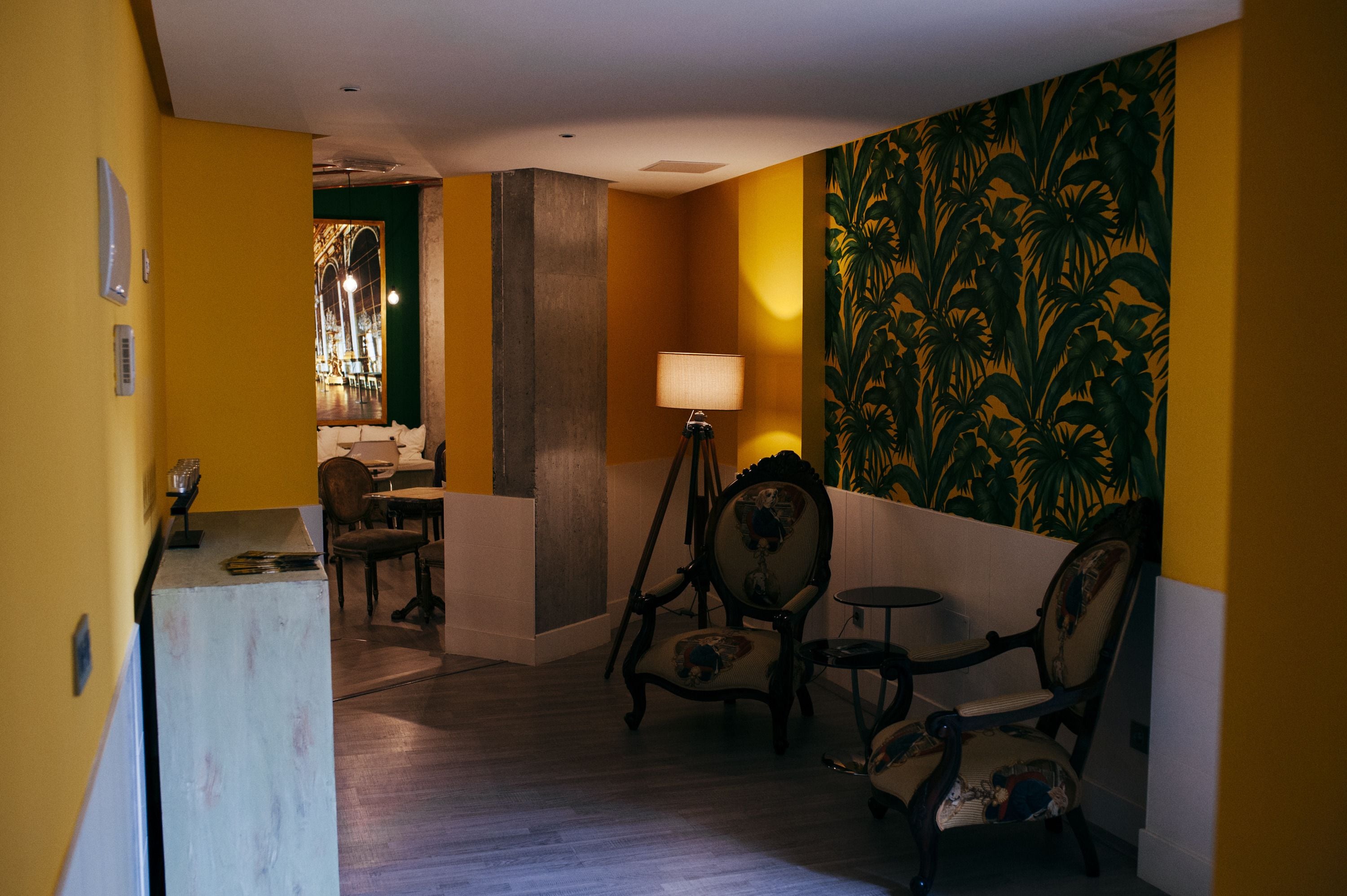 A room with yellow walls, a wooden floor, and a large mirror with a tropical wallpaper. There is a lamp on a tripod and two chairs.