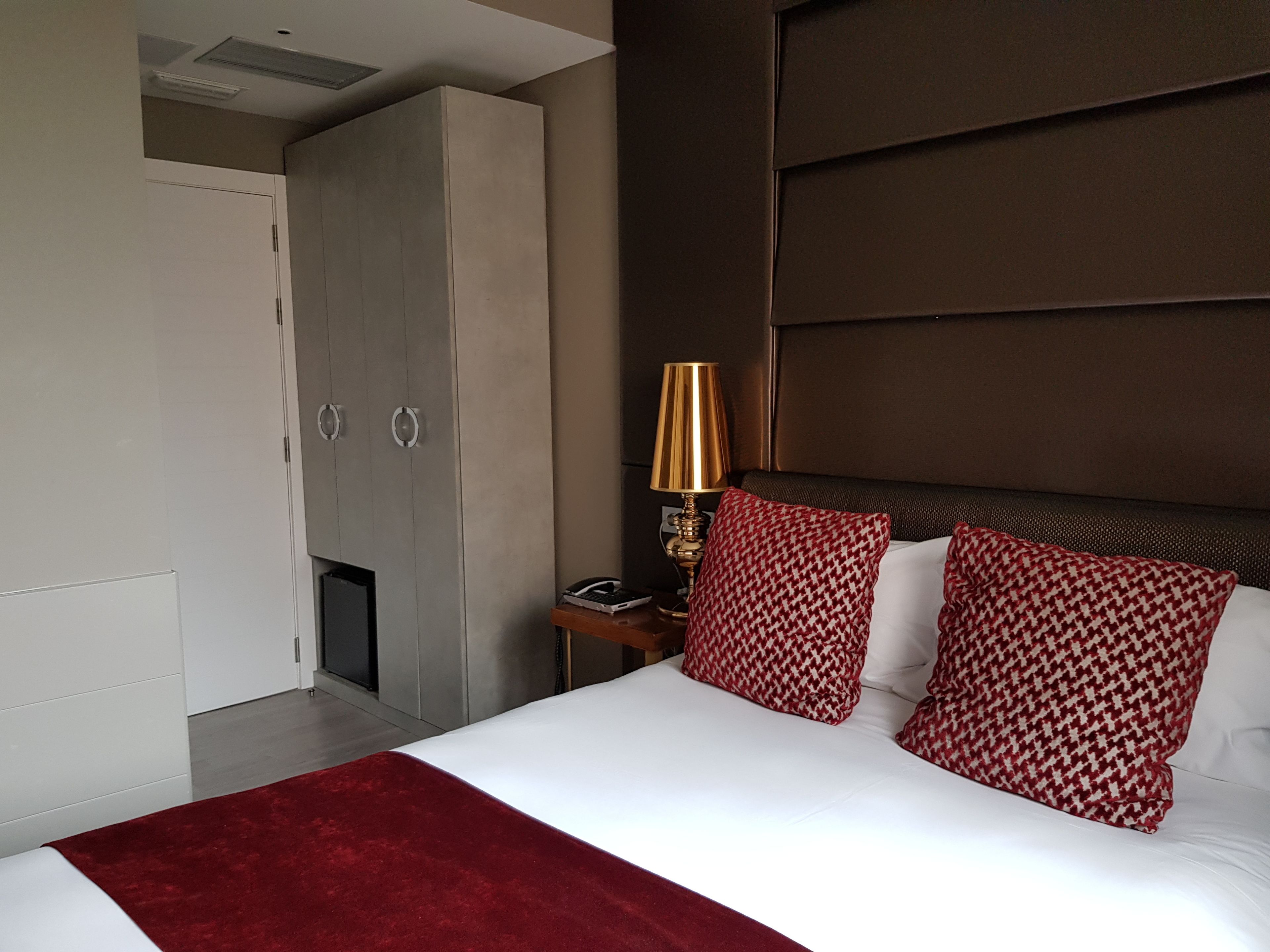 Habitación de hotel con una cama grande, un armario, una mesita de noche con lámpara y almohadones estampados.