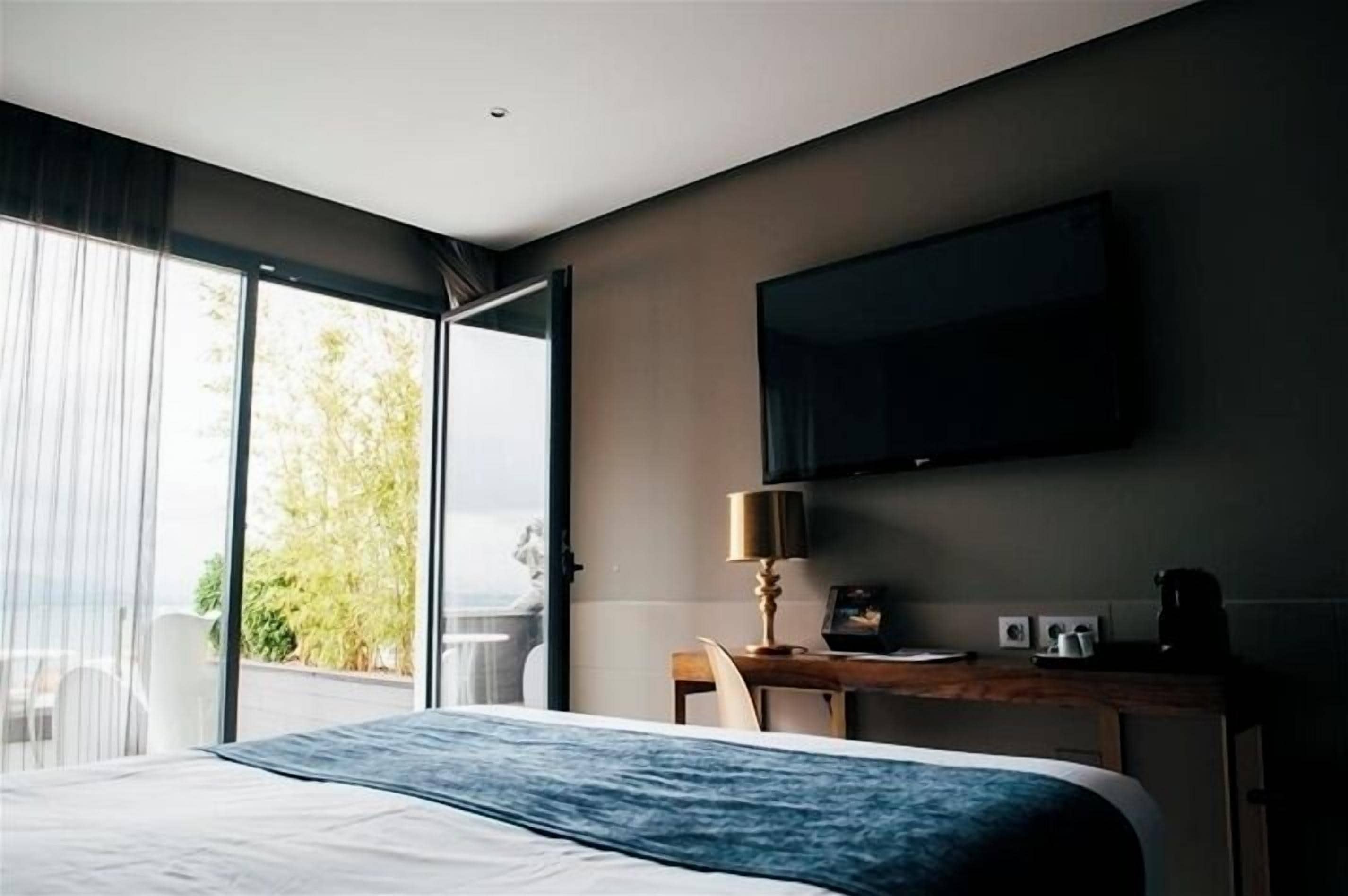 A hotel room with a bed, a desk with a lamp, a flat-screen TV, and a view of greenery outside.