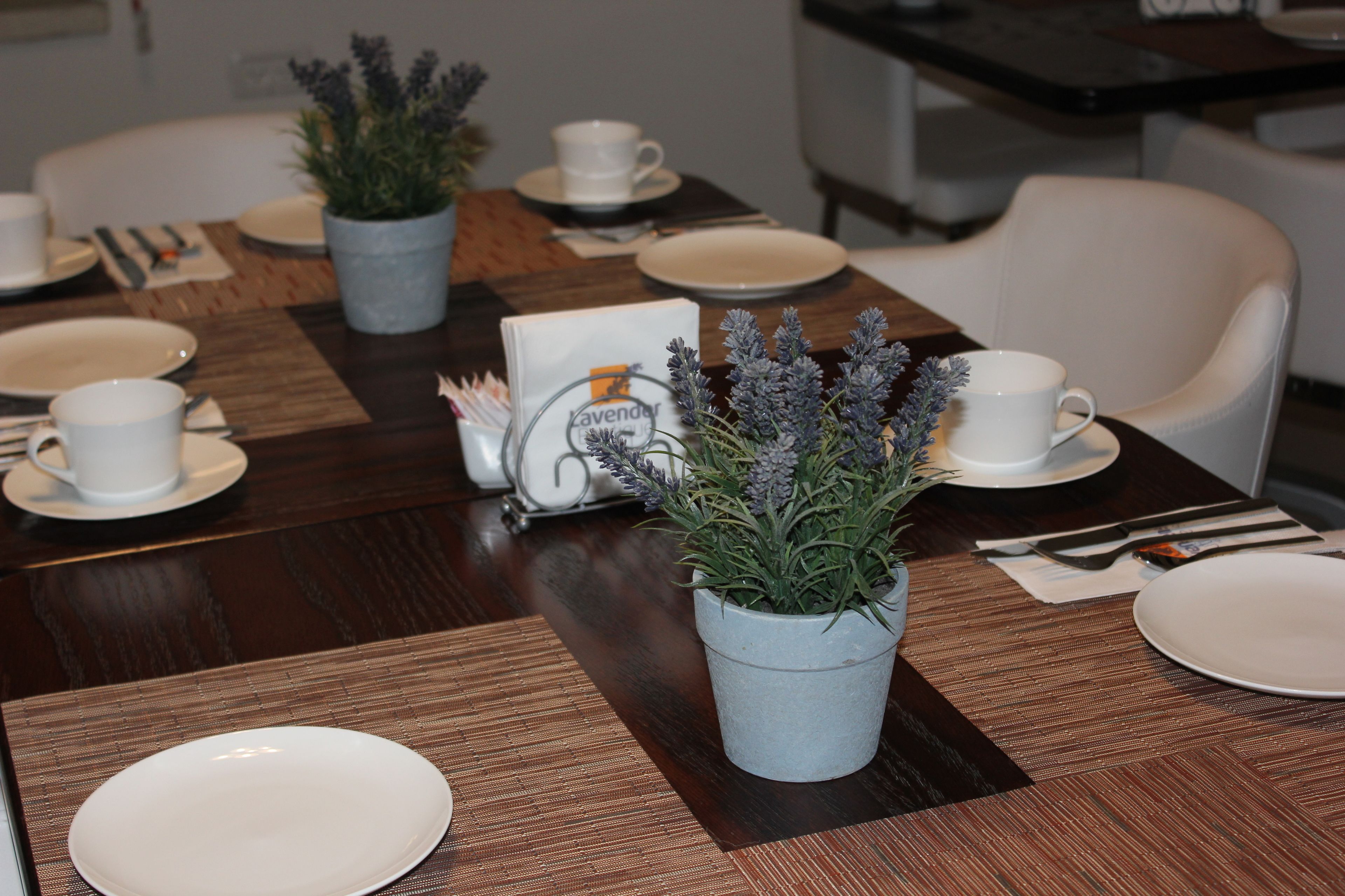 Mesa de jantar posta com pratos, xícaras e talheres brancos, com duas plantas de lavanda em vasos e um cardápio.