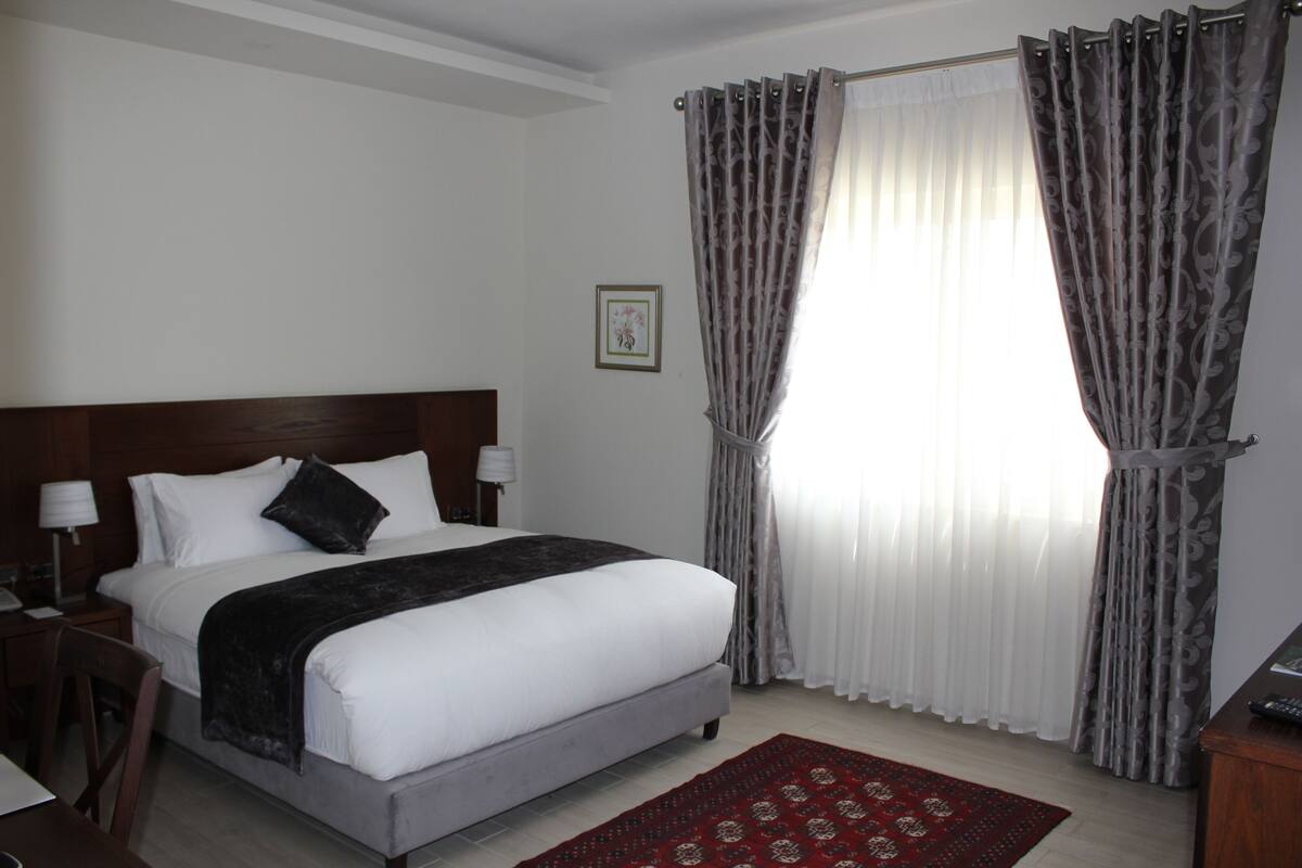A hotel room with a large bed, a nightstand, a desk, and a window with curtains.